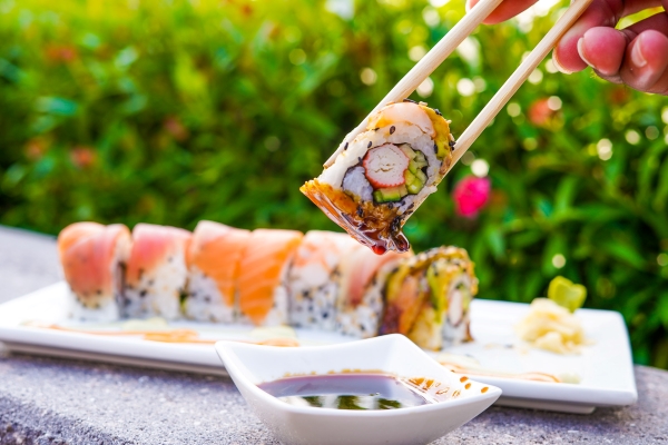 A colorful roll of sushi against a green and floral backdrop with a hand holding a roll dipped in soy sauce with chopsticks in the foreground.
