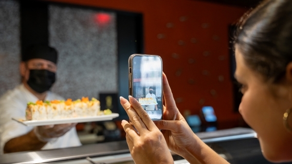 A woman taking an Instagram picture of plate of sushi held by a sushi chef.