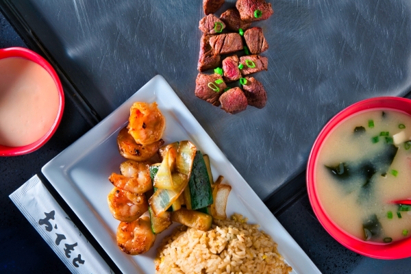 An overhead shot of a black countertop with steak, a bowl of soup, a side sauce, and a plate of shrimp, vegetables, and fried rice.