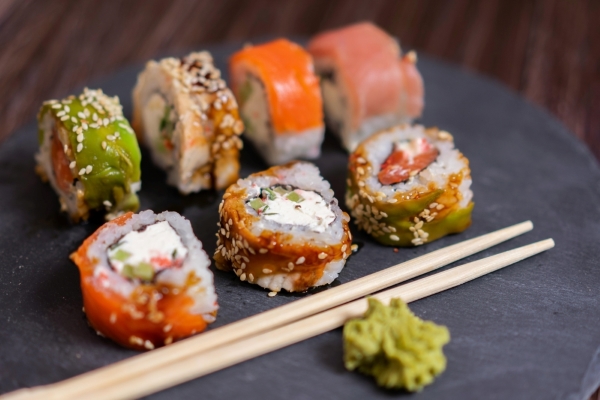 Several colorful sushi rolls filled with cream cheese on a cement plate beside a pair of wooden chopsticks and green wasabi.