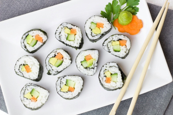 A white plate with rolls of sushi stuffed with carrot, avocado, and cucumber, with a pair of wooden chopsticks balancing on the side.