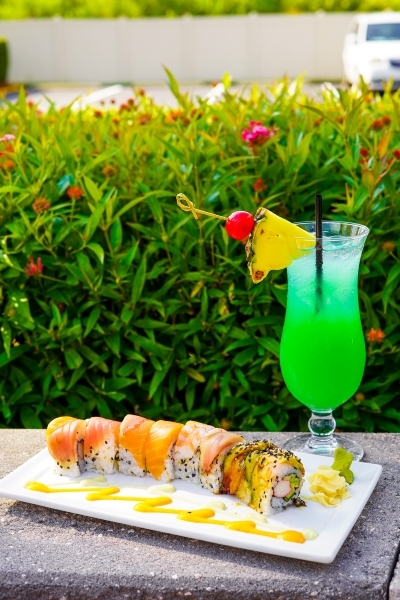 On an outdoor stone slab, a white rectangular plate with colorful sushi rolls sits beside a bright green cocktail in a hurricane glass with a pineapple garnish.  