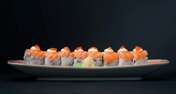 A line of sushi topped with salmon and mayo on an oblong plate against a black backdrop. Enjoy different types of sushi at Sakura Sushi. 